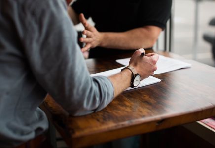Oficina de prensa y estrategia de comunicación corporativa - La imagen muestra a dos personas discutiendo sobre documentos oficiales, sentadas en una mesa de madera, con papel y bolígrafo en mano. El contexto representa la planificación de comunicados de prensa y estrategias de relaciones con los medios. Us Company Advisor ayuda a las empresas italianas a ganar visibilidad en el mercado estadounidense a través de una gestión profesional de la comunicación y las relaciones públicas.