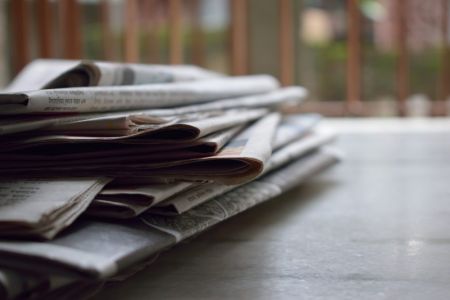 Oficina de prensa y relaciones con los medios - La imagen muestra una pila de periódicos en papel sobre un escritorio, con un fondo borroso que sugiere un entorno profesional. Esto representa la difusión de noticias, notas de prensa y gestión de la reputación corporativa. Us Company Advisor apoya a las empresas italianas en la creación y distribución de contenidos de prensa para ganar visibilidad y credibilidad en el mercado estadounidense.