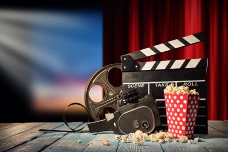 Industria del entretenimiento y el mundo del espectáculo en EE. UU. - La imagen muestra una cámara de cine antigua, un carrete de película, una claqueta del director y un contenedor de palomitas de maíz sobre una mesa de madera, con una pantalla de cine borrosa y cortinas rojas en el fondo. Esto representa el mundo del cine, la televisión y el entretenimiento. Us Company Advisor apoya a empresas y profesionales italianos en su entrada y desarrollo en el sector del entretenimiento en Estados Unidos, ofreciendo consultoría para producciones, eventos y estrategias de marketing.