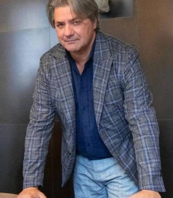 L'immagine ritrae Paolo Odierna e Lorenzo Lisi che camminano con sicurezza lungo una strada urbana. Entrambi sono vestiti con eleganza: l'uomo a sinistra indossa un completo grigio con un cappotto a quadri lungo, una sciarpa scura e scarpe lucide, mentre l'uomo a destra indossa un completo blu con una sciarpa a fantasia e scarpe nere. Lo sfondo sfocato mostra un contesto cittadino con auto parcheggiate e edifici storici, creando un'atmosfera dinamica e raffinata.