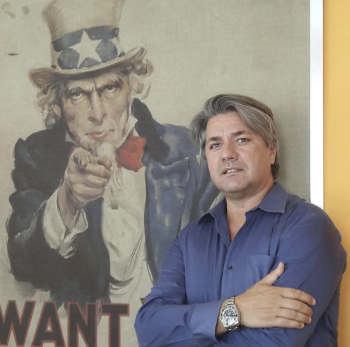 Paolo Odierna in posa davanti al celebre poster di Uncle Sam 'I Want You' - L'immagine mostra un uomo con camicia blu e orologio al polso, in piedi con le braccia incrociate, davanti al famoso manifesto di reclutamento statunitense. Questa scena simboleggia il legame con il business negli USA e l'opportunità per imprenditori italiani di espandersi nel mercato americano. Us Company Advisor offre consulenza strategica per aziende che vogliono crescere negli Stati Uniti.