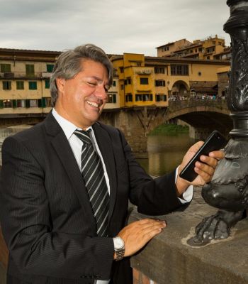 L'immagine ritrae Paolo Odierna e Lorenzo Lisi che camminano con sicurezza lungo una strada urbana. Entrambi sono vestiti con eleganza: l'uomo a sinistra indossa un completo grigio con un cappotto a quadri lungo, una sciarpa scura e scarpe lucide, mentre l'uomo a destra indossa un completo blu con una sciarpa a fantasia e scarpe nere. Lo sfondo sfocato mostra un contesto cittadino con auto parcheggiate e edifici storici, creando un'atmosfera dinamica e raffinata.