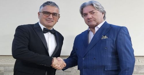 L'immagine mostra due uomini che si stringono la mano in un contesto formale. L'uomo a sinistra indossa un elegante smoking nero con papillon e occhiali, mentre l'uomo a destra è vestito con un raffinato abito blu gessato, una camicia bianca aperta senza cravatta e un fazzoletto da taschino dorato. Entrambi hanno un'espressione seria e professionale. Lo sfondo è chiaro e neutro, con dettagli architettonici che suggeriscono un ambiente di prestigio, come una sala per eventi o una location per incontri d'affari.