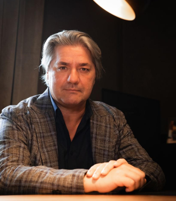 L'immagine ritrae Paolo Odierna con capelli grigi pettinati all'indietro, seduto a un tavolo in un ambiente dall'illuminazione calda e intima. Indossa una giacca a quadri su una camicia scura e ha un'espressione seria e determinata. Sopra di lui, una lampada emana una luce soffusa che accentua i contorni del volto, creando un'atmosfera elegante e sofisticata. Lo sfondo è scuro e sfocato, dando risalto alla figura in primo piano.